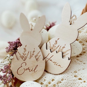 May include: Two wooden Easter bunny ornaments with the names "Emil" and "Hania" engraved on them. The bunnies are holding a wooden egg with a floral design.