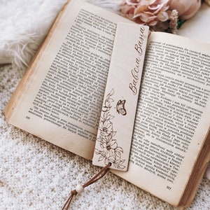 Op de afbeelding: Een houten bladwijzer met de tekst "Babcia Basia" en bloemen- en vlindergravures. De bladwijzer ligt op een open boek met verouderde pagina's, met een leren koordje eraan. Het boek is geopend op pagina 320.