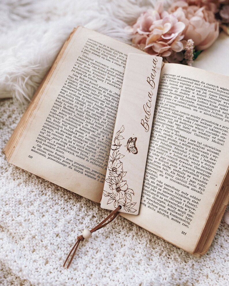 Op de afbeelding: Een houten bladwijzer met de tekst "Babcia Basia" en bloemen- en vlindergravures. De bladwijzer ligt op een open boek met verouderde pagina's, met een leren koordje eraan. Het boek is geopend op pagina 320.