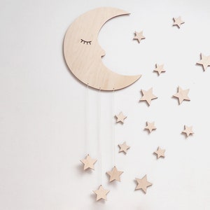 May include: A wooden crescent moon with a sleeping face and eyelashes hangs from the ceiling with strings attached to small wooden stars. The moon and stars are painted in a light wood color.