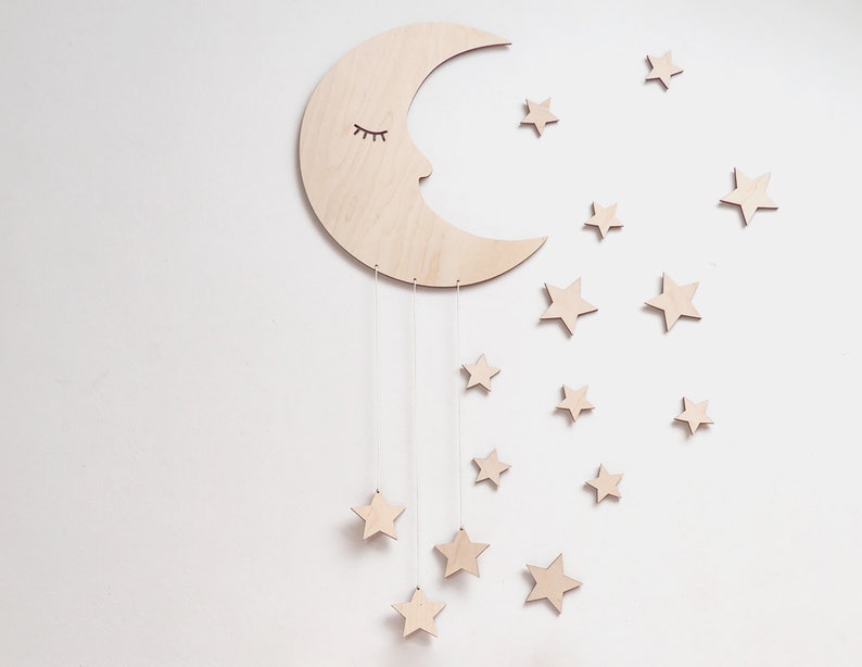 May include: A wooden crescent moon with a sleeping face and eyelashes hangs from the ceiling with strings attached to small wooden stars. The moon and stars are painted in a light wood color.