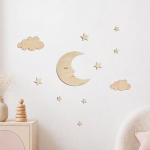 Peut inclure: Un ensemble de décorations murales en bois clair comprenant un croissant de lune avec un visage aux yeux fermés, deux formes de nuages et sept petites étoiles. Les pièces sont disposées pour créer un décor céleste sur un mur clair.