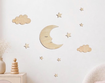 May include: A set of light-colored wooden wall decorations featuring a crescent moon with a closed-eye face, two cloud shapes, and seven small stars. The pieces are arranged to create a celestial display on a plain, light-colored wall surface.