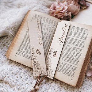 Op de afbeelding: Houten bladwijzers met bloemen- en vlindermotieven, gepersonaliseerd met de namen "Babcia Basia" en "Amelia". Ze zijn bevestigd aan een leren koord met houten kralen, op een open boek.