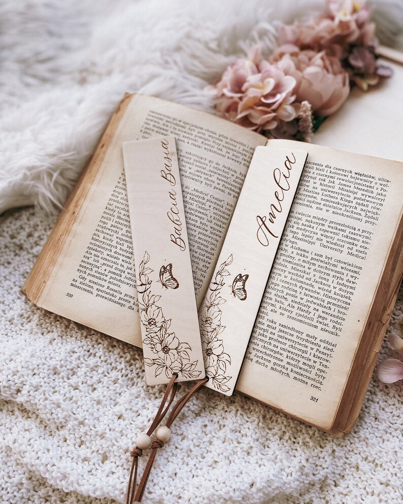 Op de afbeelding: Houten bladwijzers met bloemen- en vlindermotieven, gepersonaliseerd met de namen "Babcia Basia" en "Amelia". Ze zijn bevestigd aan een leren koord met houten kralen, op een open boek.