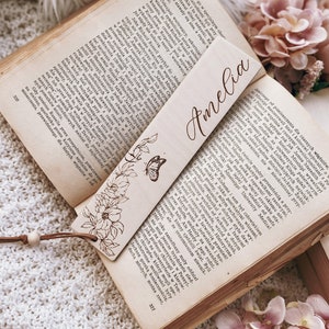 Op de afbeelding: Houten bladwijzer met de naam "Amelia" gegraveerd in een sierlijk lettertype. De bladwijzer heeft een bloemen- en vlinderontwerp. Het ligt op een open boek met verouderde pagina's.