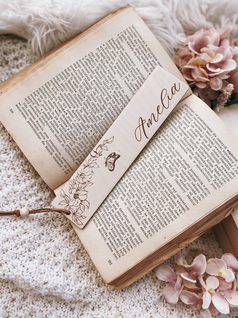 Op de afbeelding: Houten bladwijzer met de naam "Amelia" gegraveerd in een sierlijk lettertype. De bladwijzer heeft een bloemen- en vlinderontwerp. Het ligt op een open boek met verouderde pagina's.