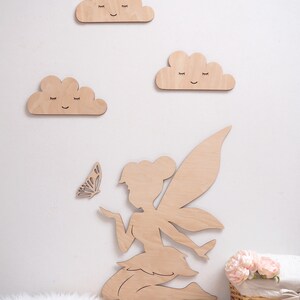 May include: Wooden fairy silhouette with wings, holding a butterfly, with three wooden clouds with smiling faces on a white wall.