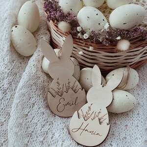 May include: Two wooden bunny-shaped Easter egg ornaments with the names "Emil" and "Hania" engraved on them. The ornaments are surrounded by white speckled Easter eggs in a wicker basket.