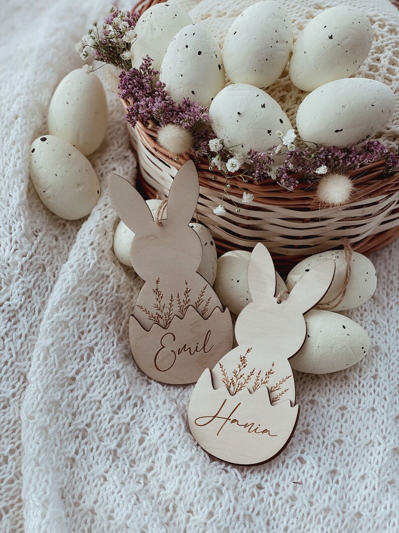 May include: Two wooden bunny-shaped Easter egg ornaments with the names "Emil" and "Hania" engraved on them. The ornaments are surrounded by white speckled Easter eggs in a wicker basket.
