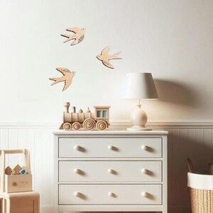 Puede incluir: Tres decoraciones de madera con forma de golondrina est&aacute;n montadas en una pared blanca. Las golondrinas son de color marr&oacute;n claro con detalles grabados. Debajo de las golondrinas hay una c&oacute;moda blanca con tres cajones y un tren de juguete de madera.