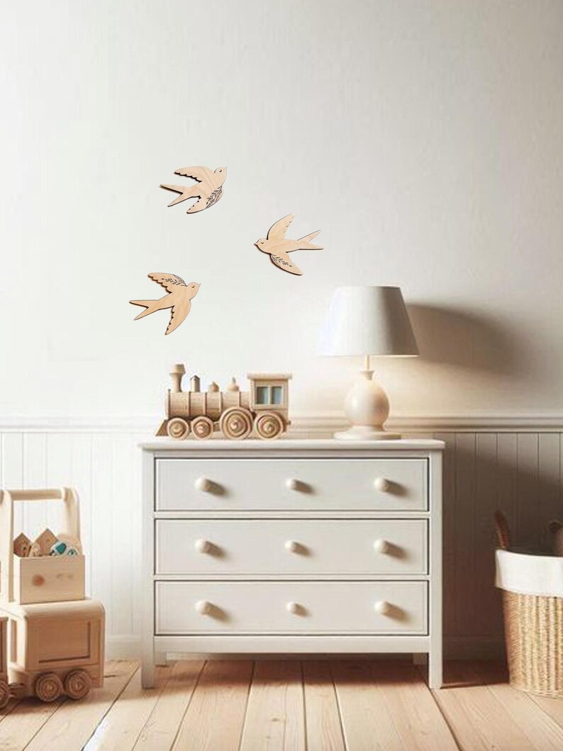Puede incluir: Tres decoraciones de madera con forma de golondrina est&aacute;n montadas en una pared blanca. Las golondrinas son de color marr&oacute;n claro con detalles grabados. Debajo de las golondrinas hay una c&oacute;moda blanca con tres cajones y un tren de juguete de madera.