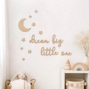 Puede incluir: Decoración de pared de madera para guardería con una luna creciente, estrellas y la frase "dream big little one". La decoración tiene un acabado de madera natural. También se ve una cesta con un conejito de peluche y un arcoíris de madera.