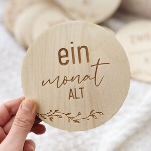 May include: A round wooden sign with the text "ein monat ALT" in a cursive font. The sign is decorated with a simple floral design around the edge.