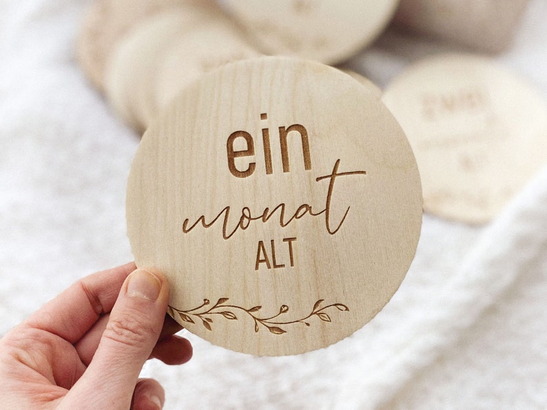 May include: A round wooden sign with the text "ein monat ALT" in a cursive font. The sign is decorated with a simple floral design around the edge.