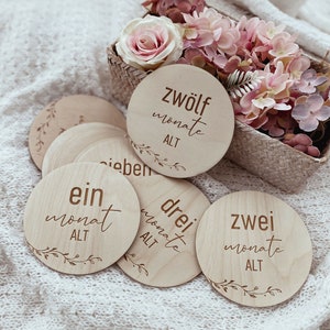 May include: Wooden baby milestone discs with the words "ein monat alt", "zwei monate alt", "drei monate alt", and "zwolf monate alt" written on them. The discs are arranged on a white knitted blanket.