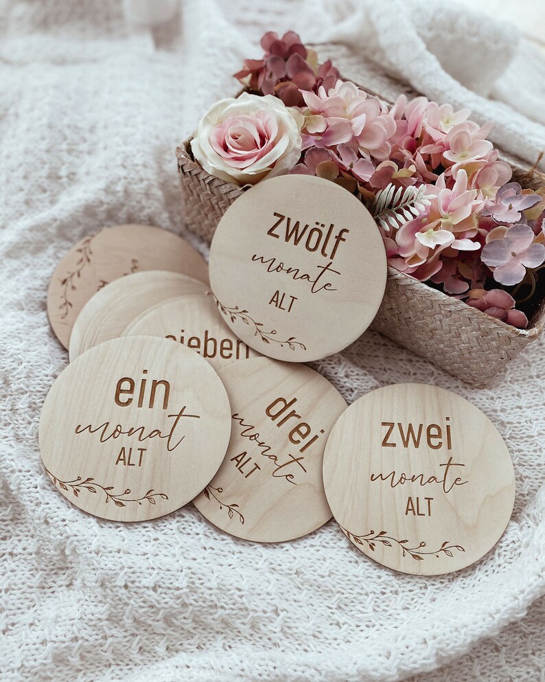 May include: Wooden baby milestone discs with the words "ein monat alt", "zwei monate alt", "drei monate alt", and "zwolf monate alt" written on them. The discs are arranged on a white knitted blanket.
