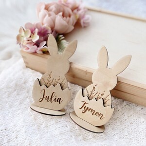 May include: Two wooden bunny place card holders with the names Julia and Tymon engraved on them. The bunnies are sitting in a cracked egg shape with a sprig of greenery.