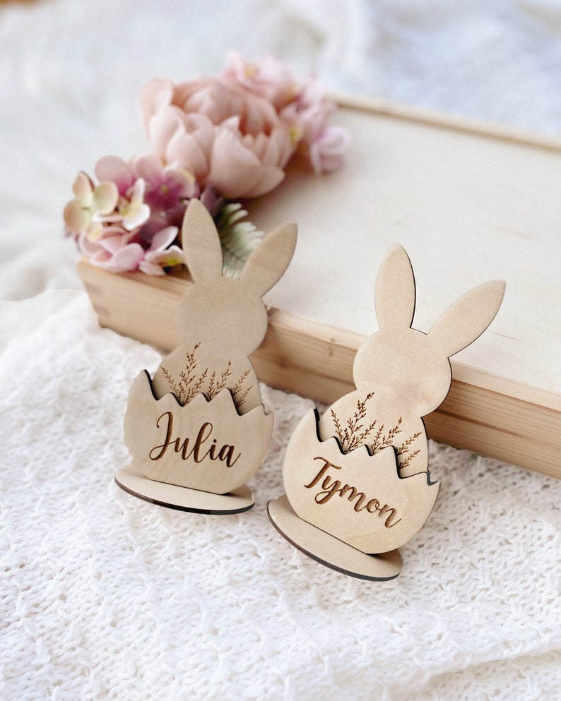 May include: Two wooden bunny place card holders with the names Julia and Tymon engraved on them. The bunnies are sitting in a cracked egg shape with a sprig of greenery.