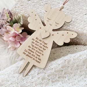 Puede incluir: Adorno de &aacute;ngel de madera con detalles de coraz&oacute;n y estrellas, con el texto en alem&aacute;n "Bin der Engel...". El &aacute;ngel tiene una cuerda para colgar y est&aacute; sobre un fondo suave y texturizado. Las alas y el cuerpo del &aacute;ngel est&aacute;n recortados en madera.
