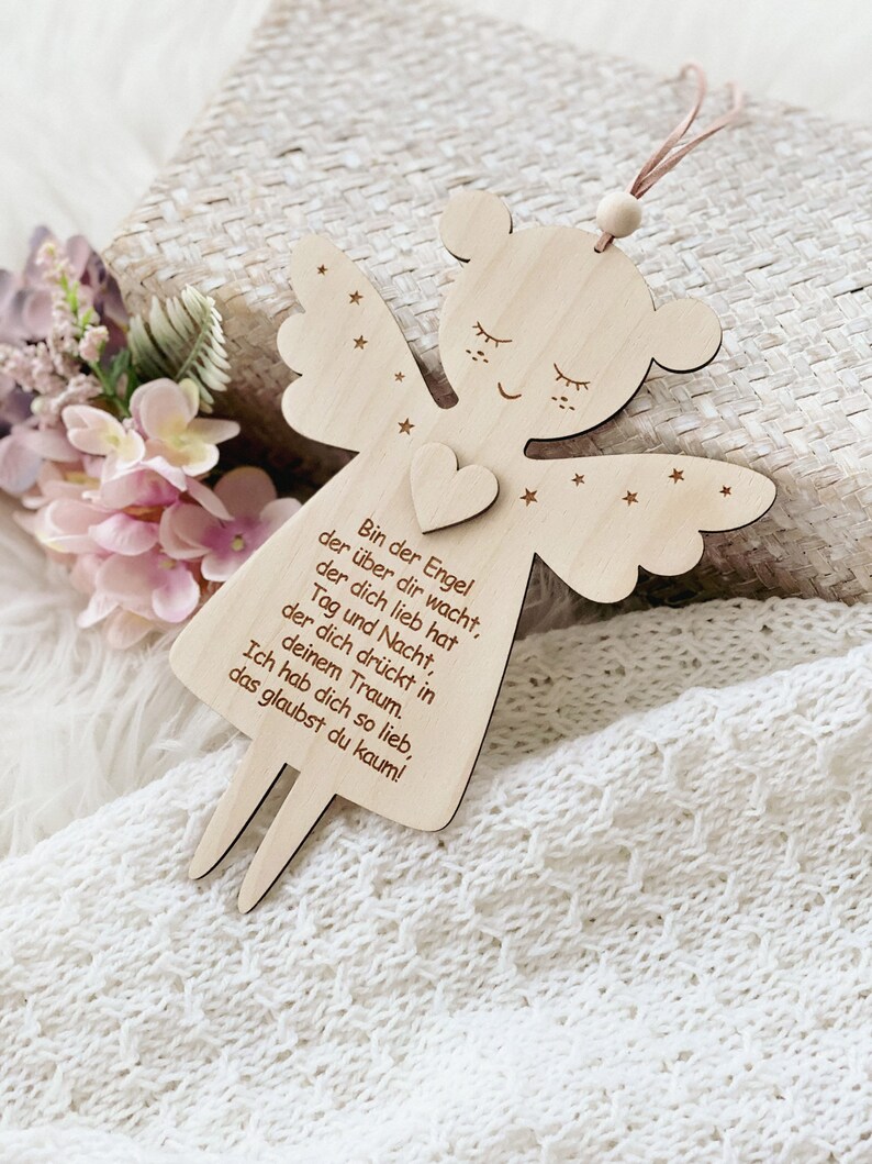 Puede incluir: Adorno de &aacute;ngel de madera con detalles de coraz&oacute;n y estrellas, con el texto en alem&aacute;n "Bin der Engel...". El &aacute;ngel tiene una cuerda para colgar y est&aacute; sobre un fondo suave y texturizado. Las alas y el cuerpo del &aacute;ngel est&aacute;n recortados en madera.