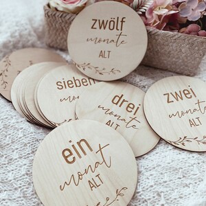 May include: A set of twelve wooden baby milestone discs with a floral design and the text "ein monat alt" (one month old), "zwei monat alt" (two months old), "drei monat alt" (three months old), "sieben monate alt" (seven months old), and "zwolf monate alt" (twelve months old).