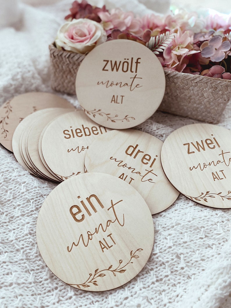 May include: A set of twelve wooden baby milestone discs with a floral design and the text "ein monat alt" (one month old), "zwei monat alt" (two months old), "drei monat alt" (three months old), "sieben monate alt" (seven months old), and "zwolf monate alt" (twelve months old).