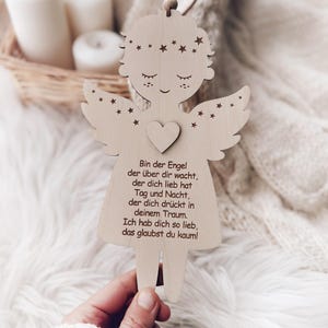 Puede incluir: Adorno de &aacute;ngel de madera con detalles de coraz&oacute;n y estrellas. El &aacute;ngel tiene los ojos cerrados y alas. El texto en el &aacute;ngel dice: "Bin der Engel der &uuml;ber dir wacht, der dich lieb hat Tag und Nacht, der dich dr&uuml;ckt in deinem Traum. Ich hab dich so lieb, das glaubst du kaum!"