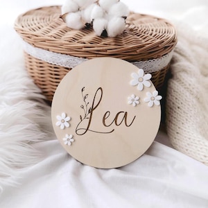 Peut inclure: Une plaque ronde en bois avec le nom "Lea" gravé en écriture cursive. La plaque est décorée de fleurs 3D blanches et d'un petit motif floral.