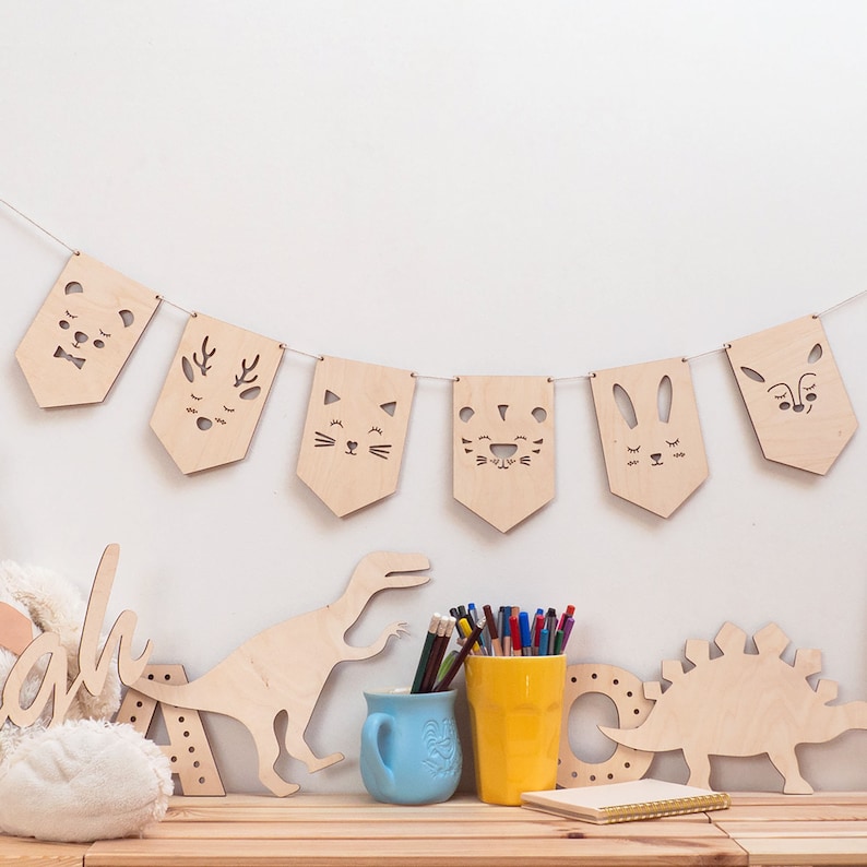 May include: A wooden banner with cut-out animal faces, including a bear, deer, cat, tiger, bunny, and fox. The banner is hanging on a white wall. There are wooden dinosaur toys and a cup of pencils on a wooden table below the banner.