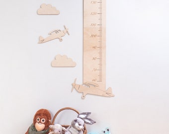 May include: Wooden growth chart with a ruler marked in inches from 60 to 130. The chart features a plane and clouds cut out of wood. The chart is mounted on a white wall.