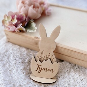 May include: Wooden Easter bunny place card holder with a brown egg shape and the name "Tymon" engraved on it. The bunny has long ears and is holding a small bouquet of flowers.