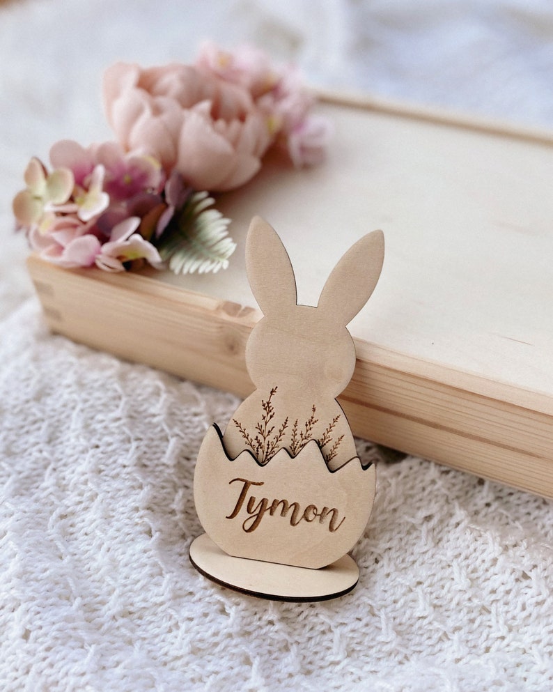 May include: Wooden Easter bunny place card holder with a brown egg shape and the name "Tymon" engraved on it. The bunny has long ears and is holding a small bouquet of flowers.