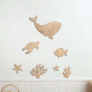Op de afbeelding: Houten wandkunstset met een walvis, schildpad, vis, zeesterren en koraal. De zeedierenfiguren zijn uit lichtgekleurd hout gesneden en op een witte muur geplaatst.