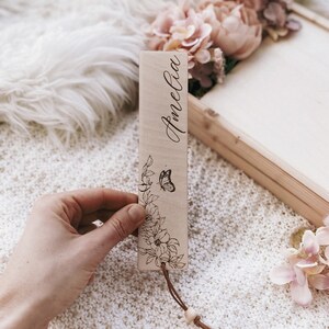 Op de afbeelding: Een houten bladwijzer met de naam "Amelia" in cursief gegraveerd. Het heeft een bloemen- en vlinderontwerp, met een leren koord en een houten kraal. De achtergrond toont een wit, getextureerd oppervlak en roze bloemen.