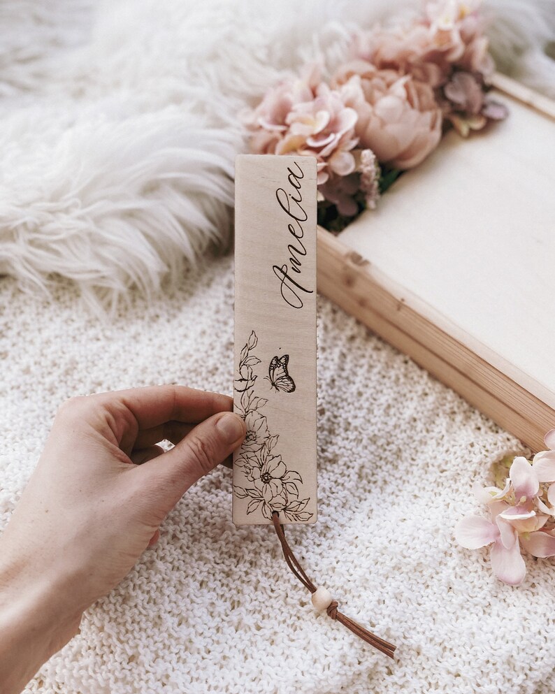 Op de afbeelding: Een houten bladwijzer met de naam "Amelia" in cursief gegraveerd. Het heeft een bloemen- en vlinderontwerp, met een leren koord en een houten kraal. De achtergrond toont een wit, getextureerd oppervlak en roze bloemen.