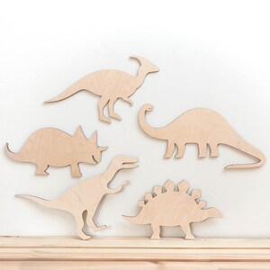 May include: Five wooden dinosaur silhouettes in various poses, including a triceratops, stegosaurus, brachiosaurus, and two others. The dinosaurs are cut from light brown wood and are arranged on a white background.