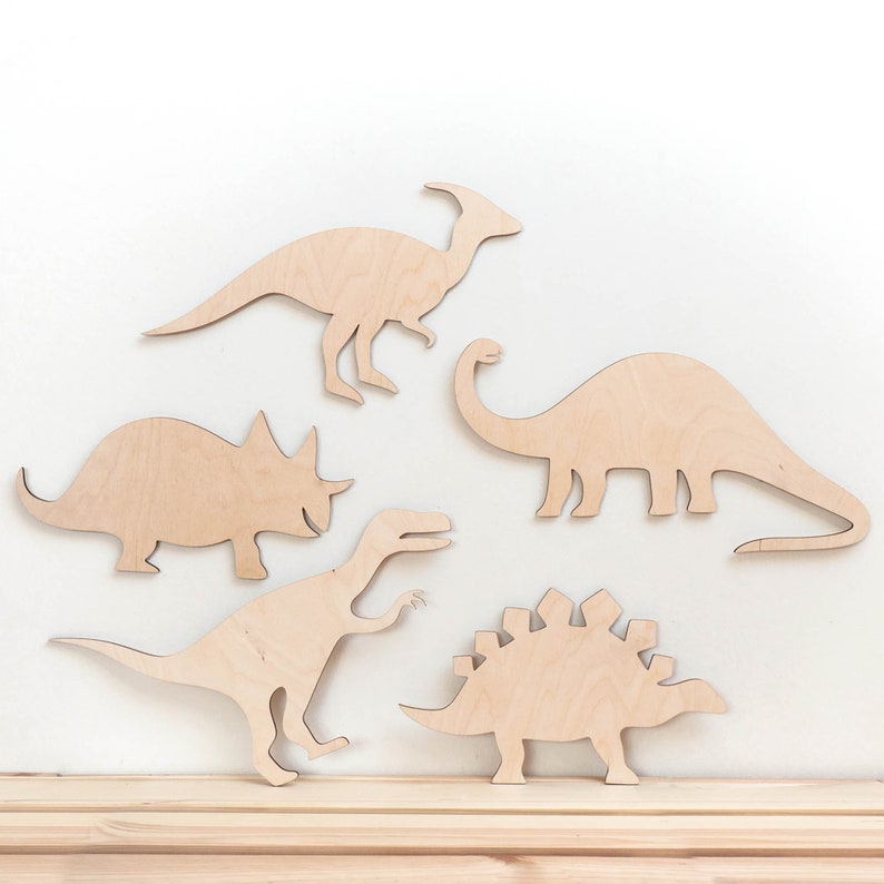 May include: Five wooden dinosaur silhouettes in various poses, including a triceratops, stegosaurus, brachiosaurus, and two others. The dinosaurs are cut from light brown wood and are arranged on a white background.