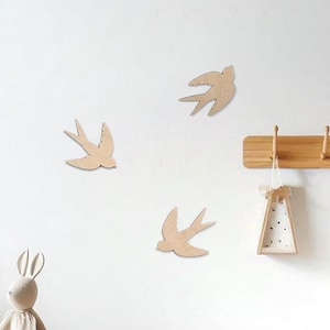 Peut inclure: Trois décorations murales en forme d'hirondelles en bois de couleur naturelle sont fixées sur un mur blanc. Les hirondelles sont dans différentes positions, donnant une impression de vol. Un porte-manteau en bois et un objet décoratif sont accrochés au mur.