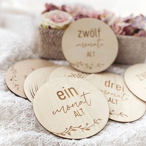 May include: A set of 12 wooden baby milestone discs with a floral design and the text "ein monat alt" (one month old) on one of the discs.