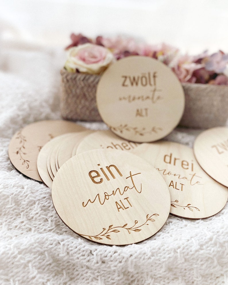 May include: A set of 12 wooden baby milestone discs with a floral design and the text "ein monat alt" (one month old) on one of the discs.