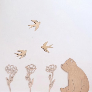 Puede incluir: Decoraci&oacute;n de pared de madera con tres p&aacute;jaros volando, tres flores y un oso. Los p&aacute;jaros tienen detalles grabados en las alas. El oso est&aacute; sentado con los ojos cerrados. Las flores tienen p&eacute;talos y tallos detallados. Todas las piezas son de madera clara.