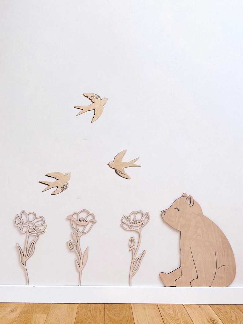 Puede incluir: Decoraci&oacute;n de pared de madera con tres p&aacute;jaros volando, tres flores y un oso. Los p&aacute;jaros tienen detalles grabados en las alas. El oso est&aacute; sentado con los ojos cerrados. Las flores tienen p&eacute;talos y tallos detallados. Todas las piezas son de madera clara.