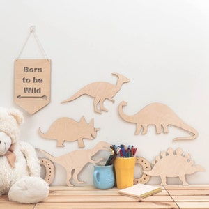 May include: Wooden dinosaur wall decor with a sign that reads "Born to be Wild". The dinosaurs are in various poses and sizes, including a triceratops, stegosaurus, and brachiosaurus. There is also a teddy bear and a cup of pencils in the image.
