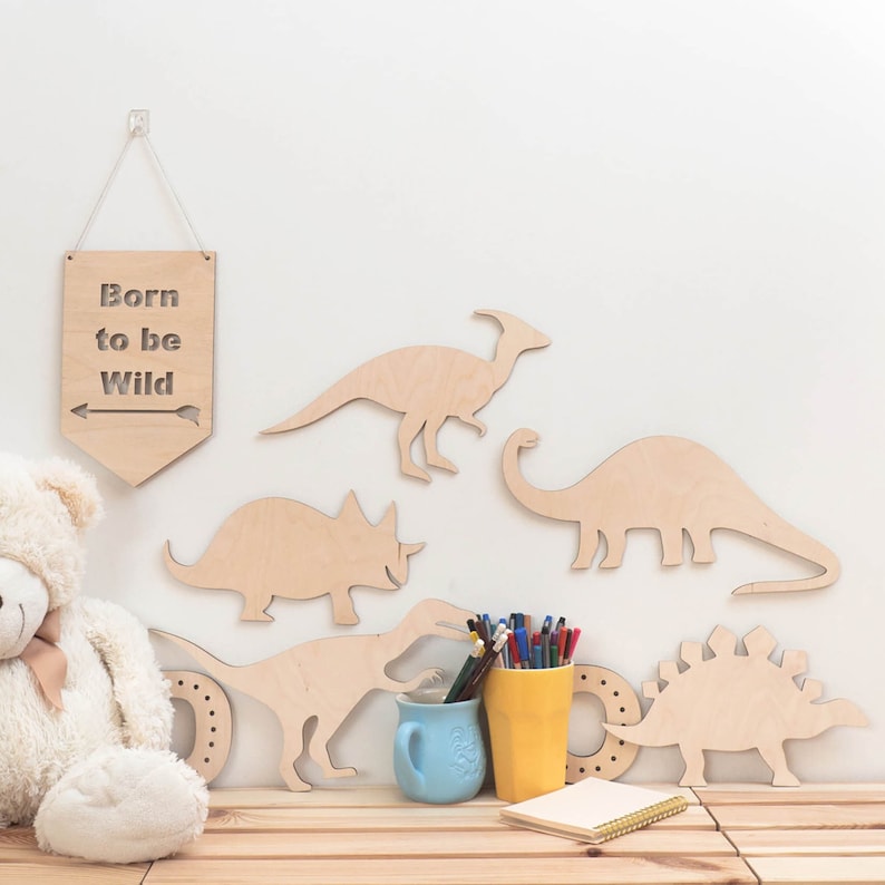 May include: Wooden dinosaur wall decor with a sign that reads "Born to be Wild". The dinosaurs are in various poses and sizes, including a triceratops, stegosaurus, and brachiosaurus. There is also a teddy bear and a cup of pencils in the image.