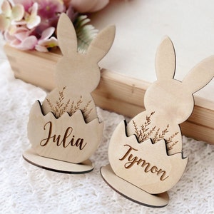 May include: Two wooden bunny place card holders with the names Julia and Tymon engraved on them. Each bunny is holding an egg with a floral design.