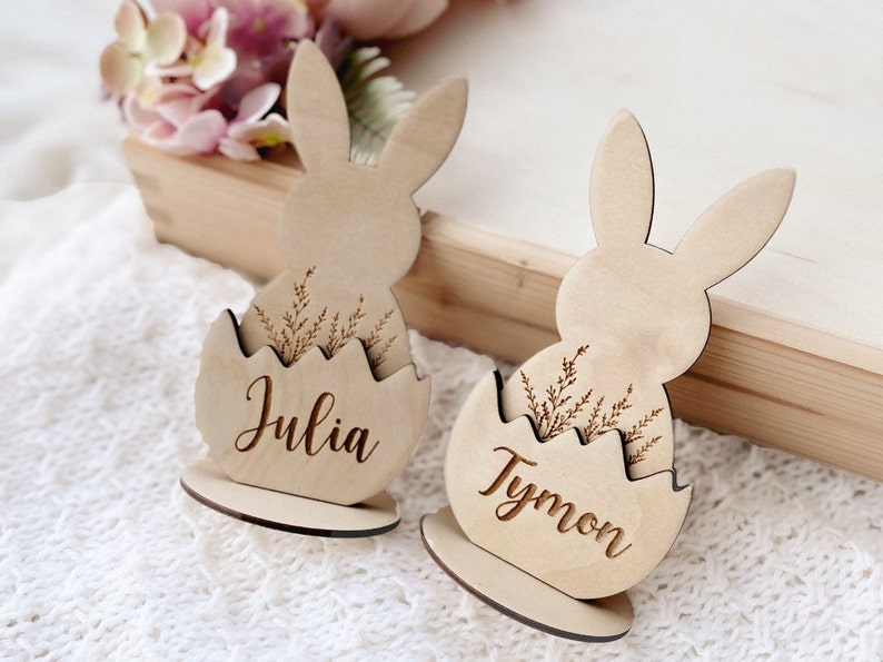 May include: Two wooden bunny place card holders with the names Julia and Tymon engraved on them. Each bunny is holding an egg with a floral design.