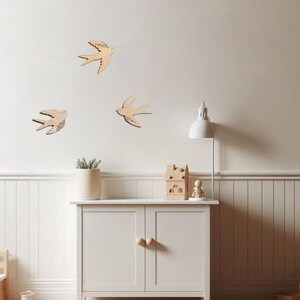 Puede incluir: Decoraci&oacute;n mural de madera con golondrinas en una habitaci&oacute;n infantil. Tres golondrinas est&aacute;n montadas en una pared blanca. Un armario blanco con pomos de madera contiene una planta en maceta, una casa de madera y una peque&ntilde;a figura. Un tren de madera est&aacute; en el suelo.
