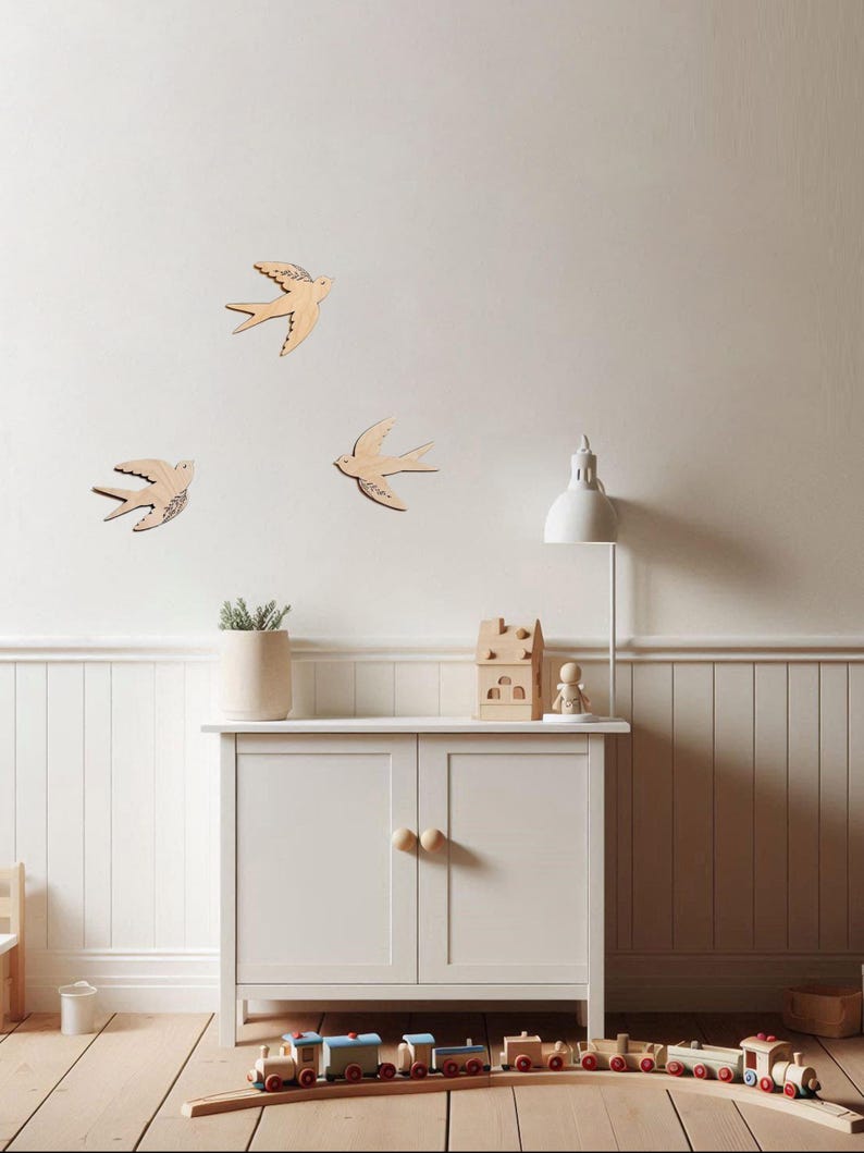 Puede incluir: Decoraci&oacute;n mural de madera con golondrinas en una habitaci&oacute;n infantil. Tres golondrinas est&aacute;n montadas en una pared blanca. Un armario blanco con pomos de madera contiene una planta en maceta, una casa de madera y una peque&ntilde;a figura. Un tren de madera est&aacute; en el suelo.