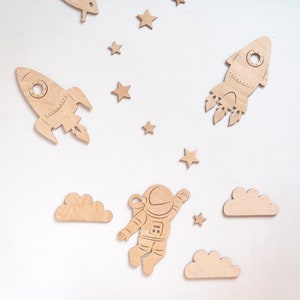 May include: Wooden astronaut, rocket, and cloud shapes in a space theme. The astronaut is in a space suit with a helmet and is floating in the air. The rocket is pointing upwards with flames coming out of the bottom. The clouds are white and fluffy.